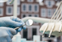 Een close-up van steriele handschoenen van een tandarts of kaakchirurg die een klein, reflecterend tandheelkundig instrument vasthouden. De setting is een zeer schone, modern verlichte tandartspraktijk. De achtergrond is extreem onscherp en toont de daken en gebouwen langs een typisch Nederlands grachtenpand, wat de locatie in Nederland aangeeft. De scène is helder en focust op professionele zorg.