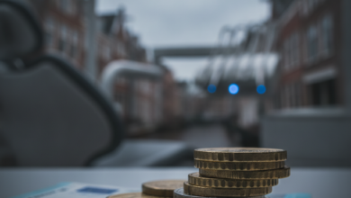 Afbeelding van euromunten en een bankbiljet op een oppervlak, met op de achtergrond een onscherpe tandartspraktijk en Nederlandse stadssilhouet, die de hoge en deels onvergoede kosten van tandheelkundige zorg symboliseert.