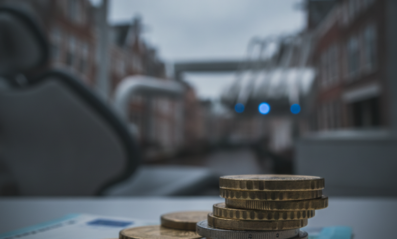 Afbeelding van euromunten en een bankbiljet op een oppervlak, met op de achtergrond een onscherpe tandartspraktijk en Nederlandse stadssilhouet, die de hoge en deels onvergoede kosten van tandheelkundige zorg symboliseert.